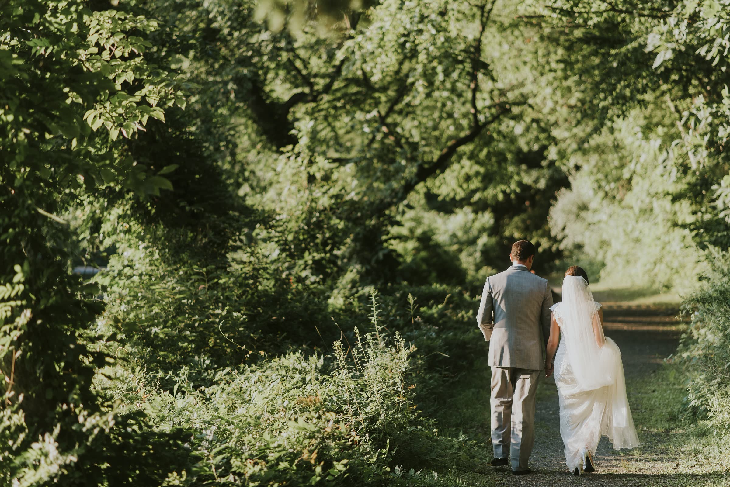 A bride and groom walking away hand in hand down a tree-lined green lane in evening light